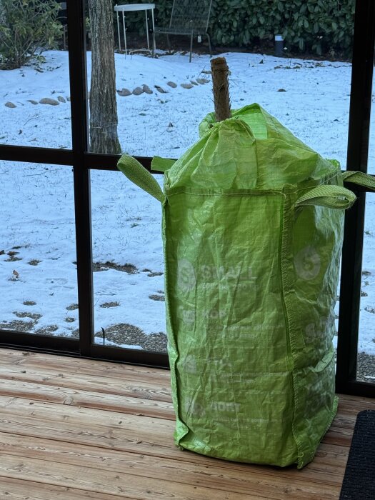 Grön förvaringssäck inomhus, med ett träföremål inuti, bredvid ett fönster som visar en snötäckt trädgård.