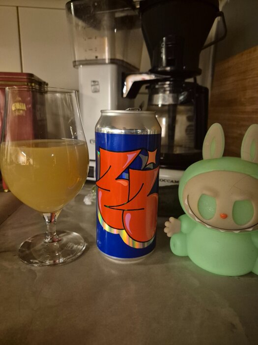Ett glas med gul dryck, en färgglad burk och en grön leksaksfigur på en köksbänk, med en kaffebryggare i bakgrunden.