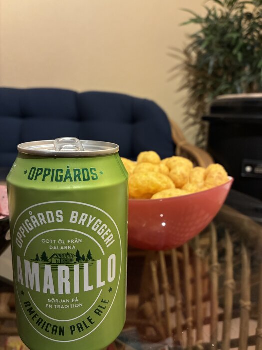 En grön burk Oppigårds Amarillo Pale Ale står på ett bord bredvid en skål med ostbågar, med en soffa och växt i bakgrunden.