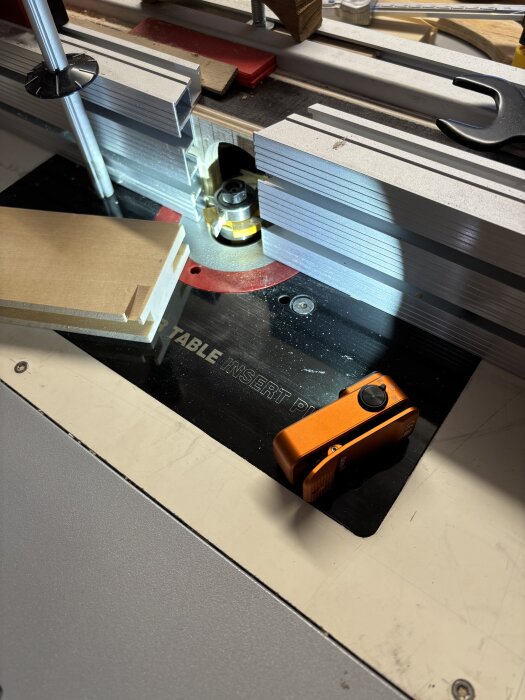 Fräsbord med träbit, frässtål och orange märkinstrument, tillsammans med stödjande metalldelar och skiftnyckel.