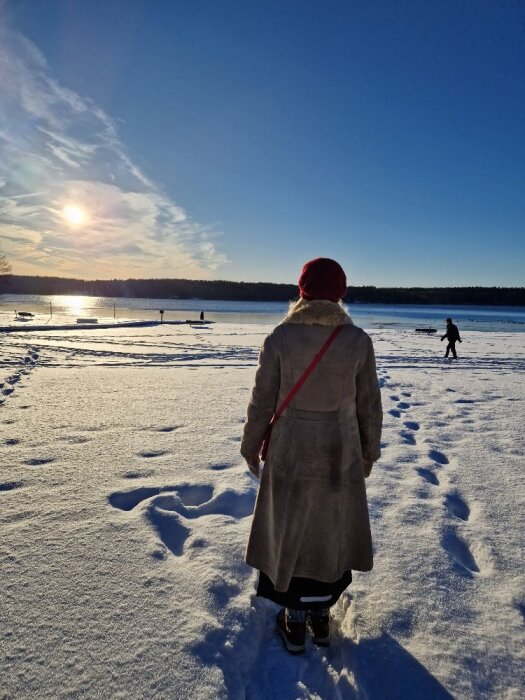 En person i vinterkläder går på snötäckt mark vid en sjö, med solen lågt på himlen och en annan person syns längre bort.