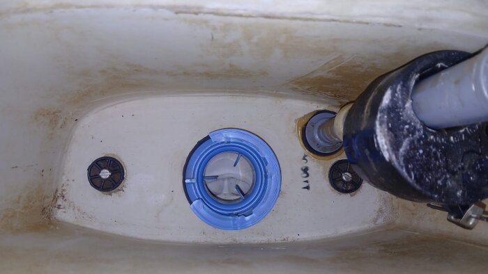 Inuti en toalettcistern med en blå plastdel fastsatt på botten, omgiven av rör och smutsavlagringar.