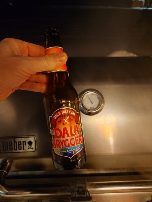 En hand håller en flaska Dala Bryggeri IPA framför en Weber-grill.