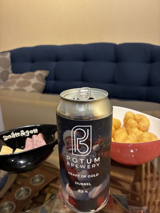 En ölburk med jultomten på etiketten står på ett bord framför en blå soffa. Bredvid finns en skål med småplock och en skål med snacks.