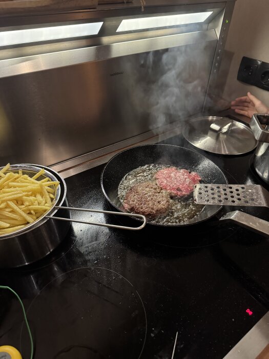 Stekpanna med wagu-burgare på spisen, medan pommes frites ligger i en kastrull bredvid.
