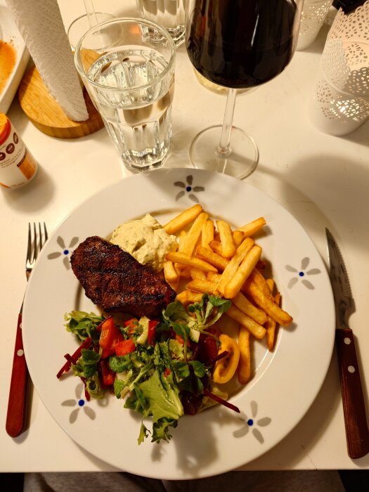 Tallrik med grillad rostas, pommes frites, sallad och sås. Glas med vin och vatten bredvid.