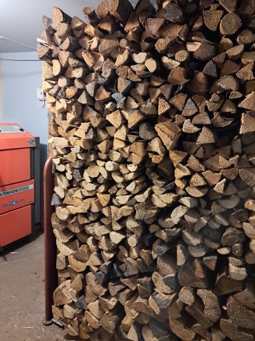 Stapel av vedträn inomhus, bredvid en röd vedpanna.