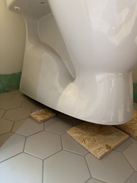 Golvstående toalettstol installerad men upplyft från golvet med träbitar, på hexagonformade kakelplattor.