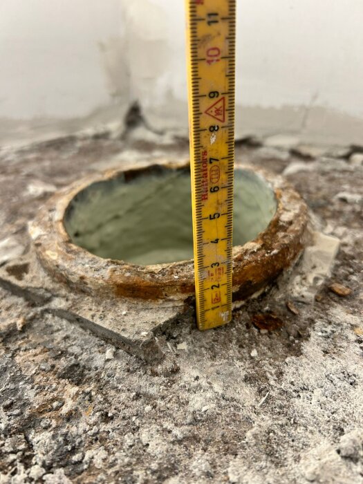 Mäthållare placerad vid blekt avloppsrör i betonggolv, visar höjd cirka 2 cm under ytan, omgivet av grov och sprucken betong.