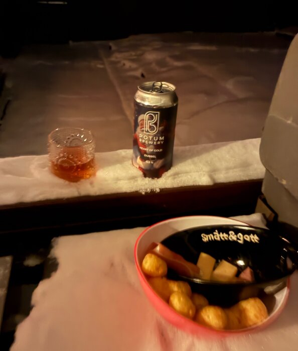Burk öl och ett glas dryck står på en snötäckt yta, bredvid en skål med snacks och ett fat med texten "smått & gott".