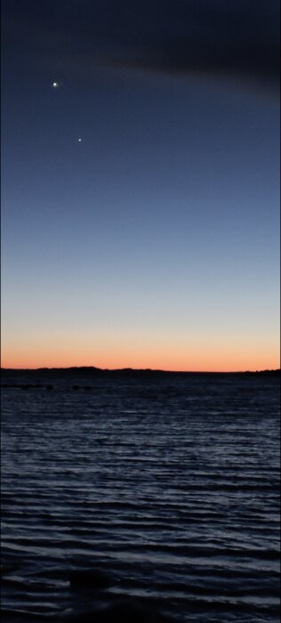 Solnedgång vid havet med två lysande himlakroppar på natthimlen över en mörk horisont och vattnet som reflekterar ljuset.