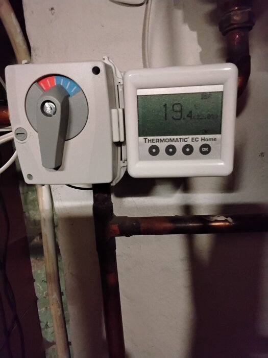 Termostat med digital display som visar 19,4 grader, inställd på uppvärmning. Monterad på vägg med kopparrör i bakgrunden.
