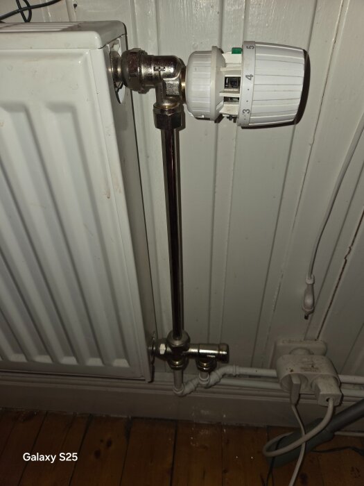 Radiator med termostatventil monterad på ett rör, omgiven av kopparrör och eluttag, fotograferad inomhus.