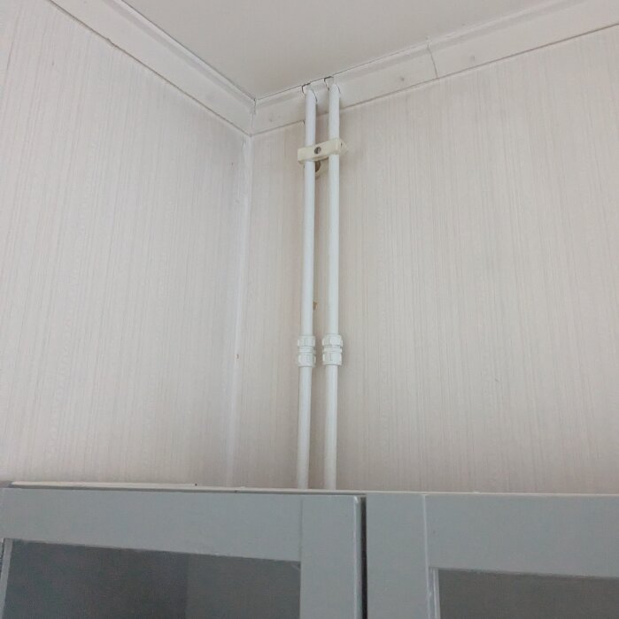 Två vertikala rör installerade längs en vägg ovanför en garderob, med beige väggbeklädnad och vit taklist.