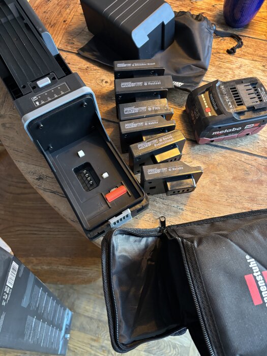 Flertal batterier och laddningsstation på ett träbord, inklusive märken som Bosch och Metabo samt en verktygsväska med blixtlås.