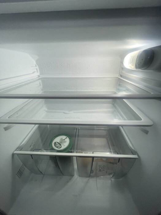 Inre av ett kylskåp med glasplan och två lådor. Ett föremål med grönt lock syns i ena lådan, och en förpackning med text syns i den andra.
