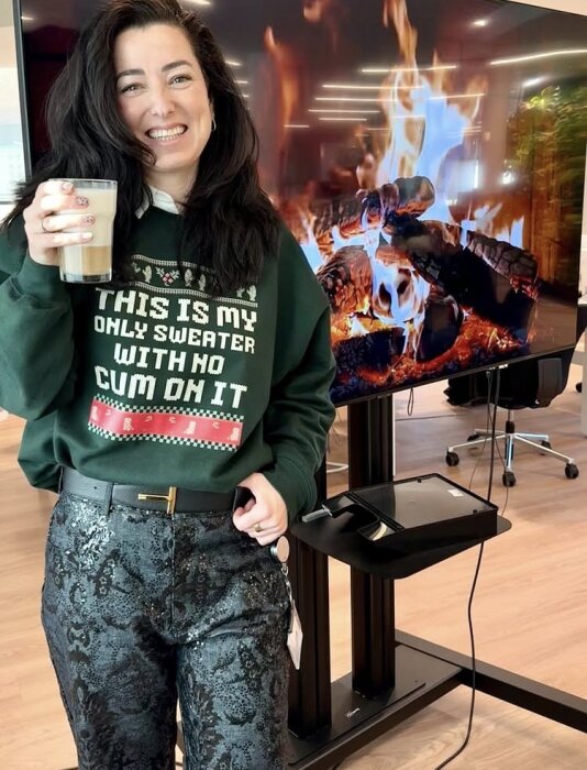 A person står med en mugg framför en TV som visar en realistisk brasa.