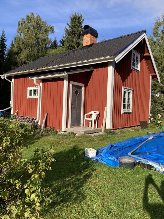 Rött trähus med svart tak och skorsten, vit plaströr och blå presenning i trädgård, solig dag, grönska i bakgrunden.