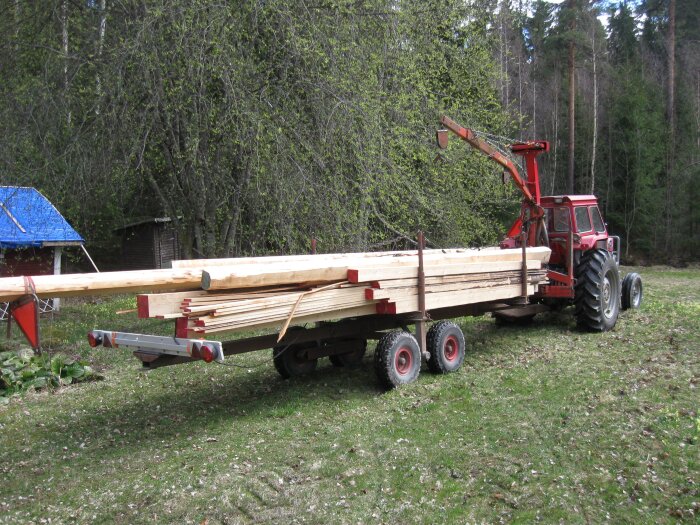Traktor med släp fullastat med långa timmerstockar, stående på en gräsmatta omgiven av skog.