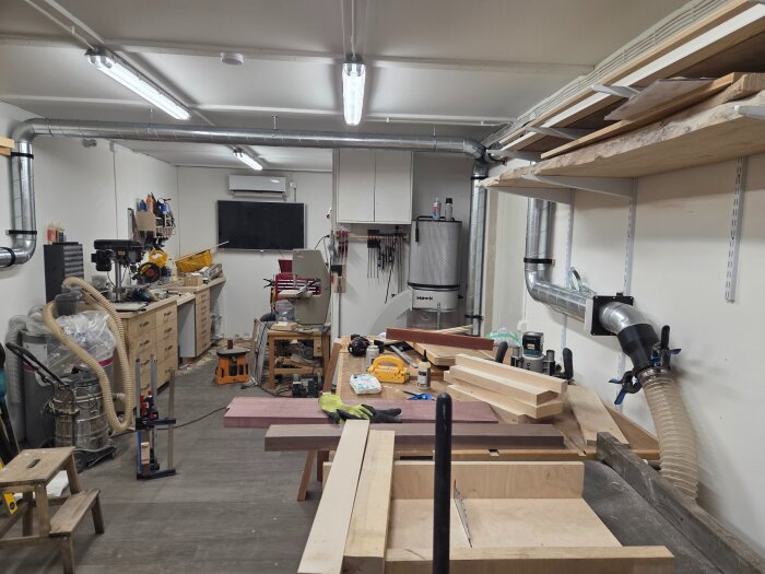 Verkstad med arbetsbänkar, hyllor och ventilationsrör. Verktyg och träbitar syns, ordnat arbetsflöde med spånrör och 3-fasuttag.