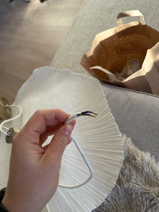 Hand som håller i avskalad lampsladd mot en vit lampskärm, papperkasse syns i bakgrunden på en soffa.