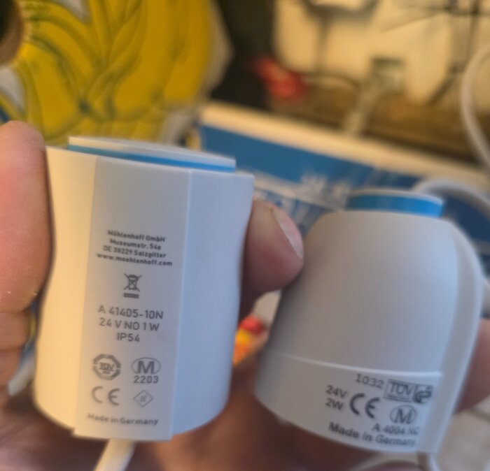 Två ställdon hålls i handen, med märkningar som visar 24V NO och NC, samt tillverkarens uppgifter och certifieringar synliga på enheten.