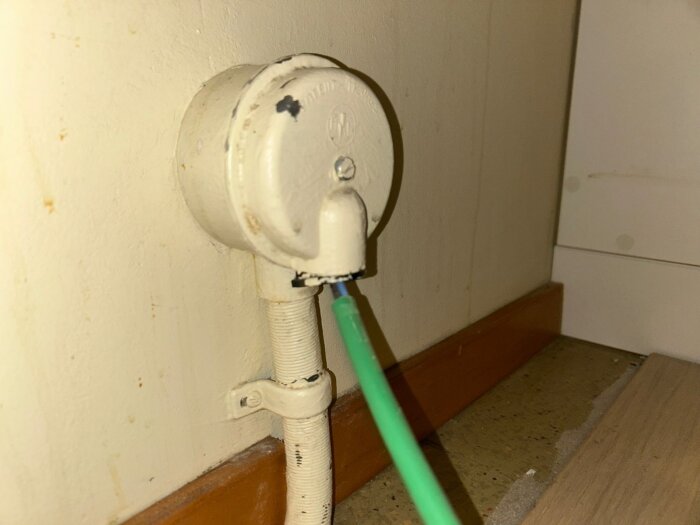 Vit spisdosa på vägg med grön kabel och skruv i locket, nära golvlist.