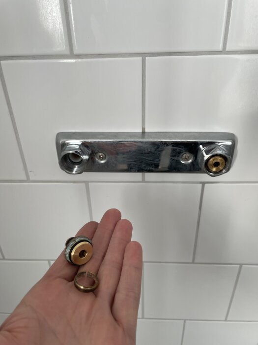 Hand som håller duschblandarens delar framför väggmonterad armatur på vit kakelvägg.