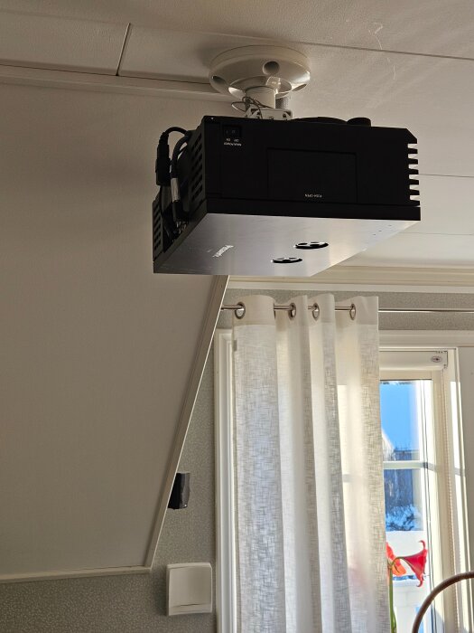 Svart bioprojektor monterad i taket med vinkeladaptrar för anslutningskablar. Den är placerad nära ett fönster med vita gardiner.