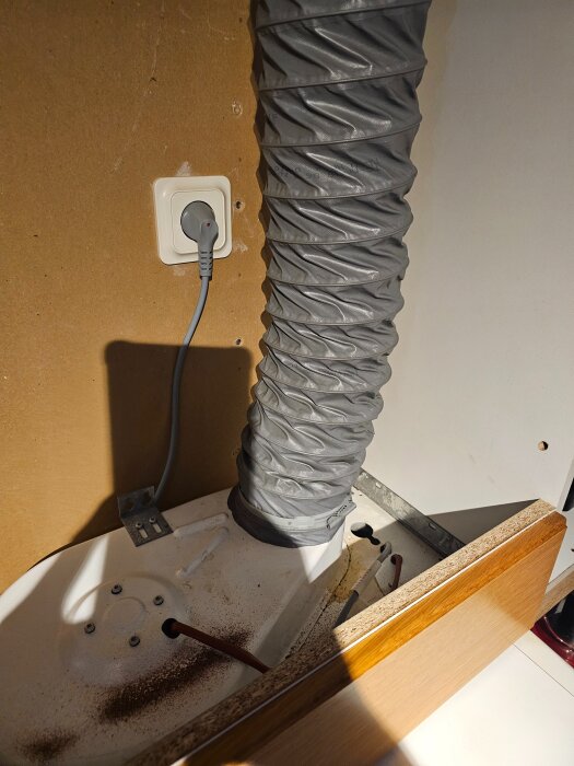 Grått ventilationsrör fäst vid köksfläkt, strömkabel ansluten till vägguttag, träpanelskant synlig, belyst under överskåp.