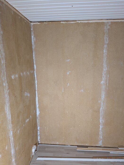 Väggar med avlägsnad tapet visar spånskivor, med rester av gammalt spackel och lister på golvet, i ett fritidshus under renovering.