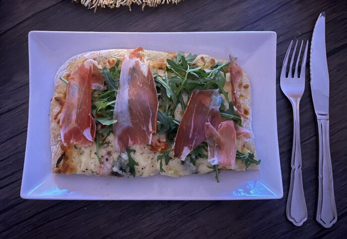 Pizza med mozzarella, ruccola och serrano på en vit tallrik. Köttkniv och gaffel ligger bredvid.
