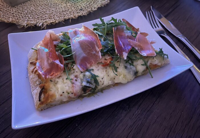 Pizza med mozzarella, ruccola och serranoskinka på en tallrik, serverad på ett bord med bestick vid sidan.