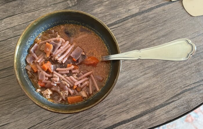 Skål med soppa innehållande rödkål, kycklingfärs, morot, paprika och pasta på ett träbord, med en sked i skålen.
