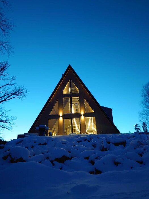 A-frame hus med takfönster och snötäckt mark, upplyst invändigt på kvällen.