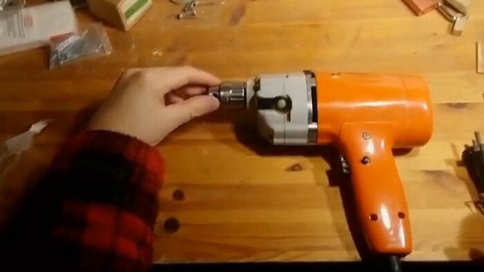 Hand justerar chucken på en orange borrmaskin på ett träbord.
