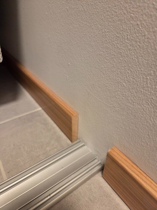 Trälist och golvsockel möter en kantlist av metall vid ett hörn på ett klinkergolv.