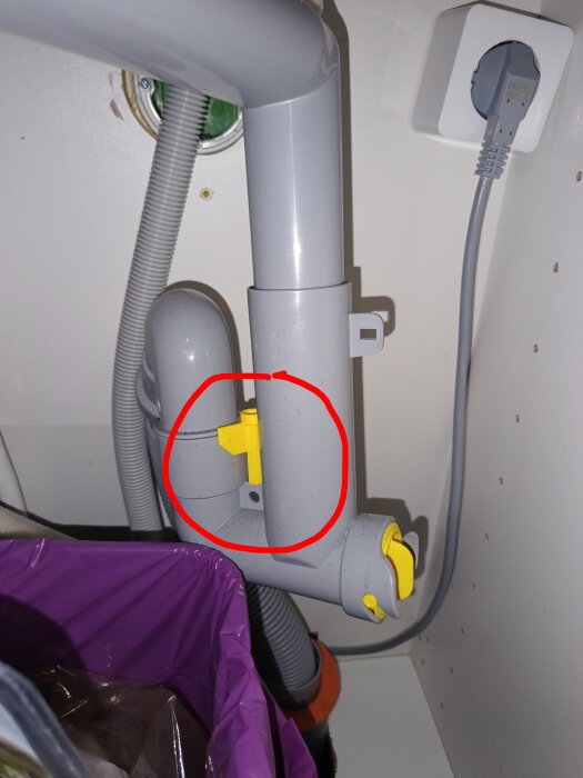 Vattenlås med gul plastnyckel från Ikea, placerad på en grå krök under ett handfat, med omgivande rör och elkabel.