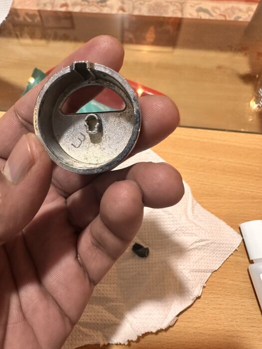 En hand som håller en metallisk, cirkulär del med en liten skåra och hål i mitten, liggandes på ett träbord med pappersservett.