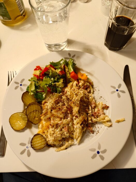 Tallrik med skinkfrestelse, sallad och inlagd gurka. Bredvid står ett glas med vatten och ett glas med svart dryck.
