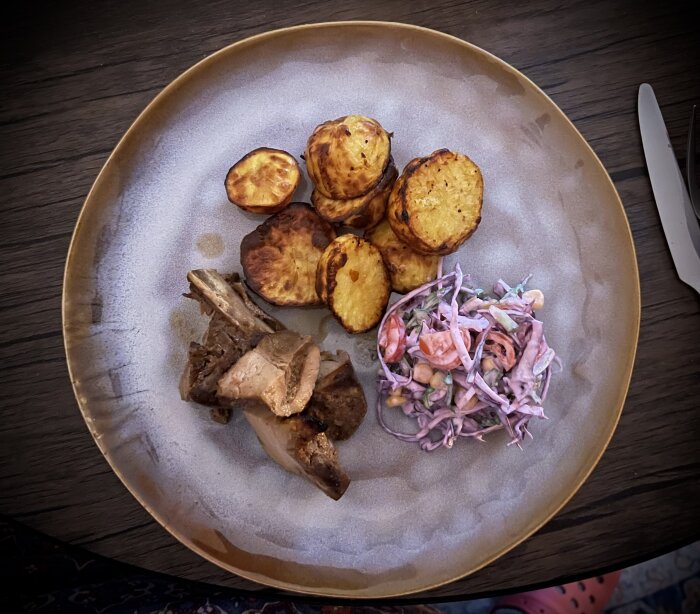 Tallrik med grillat kött, ugnsrostad potatis och coleslaw-sallad, sedd ovanifrån på ett träbord.