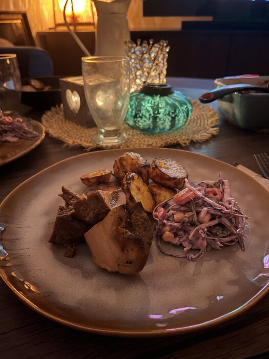 Tallrik med stekt kött, rostad potatis och coleslaw, på ett dukat bord med glas och dekorativa föremål i bakgrunden.