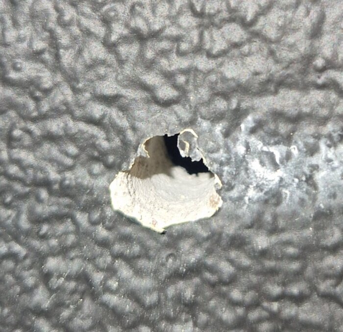 Närbild av ett hål i en grå, ojämnt strukturerad yta, där materialet runt hålet verkar vara av skadat eller sönderslaget murbruk.