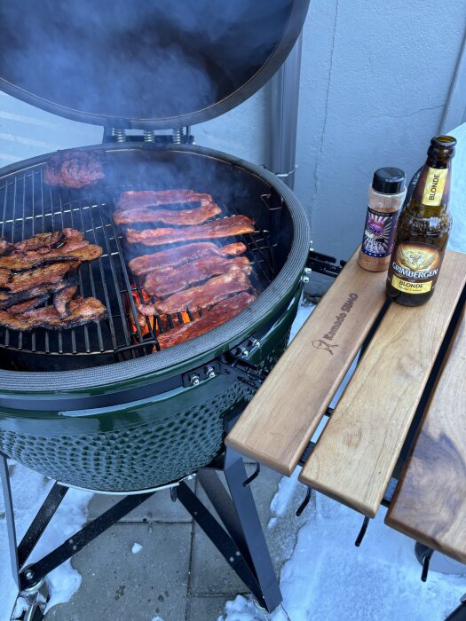 Grill med revben och köttbitar under rök. Vid sidan står en flaska öl och en kryddburk på ett träbord. Snö syns på marken.