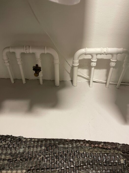 Vita rör med strypventil under en radiator, visar rördragning för flödesbegränsning i värmesystem.