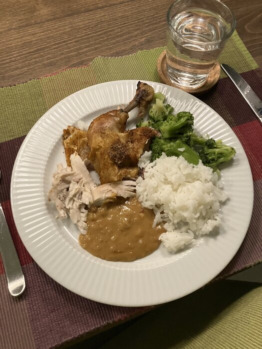 Tallrik med kyckling, ris, broccoli och sås, bredvid ett glas vatten på bordet.