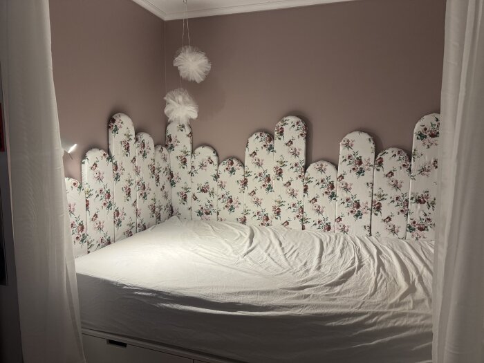 Blommiga väggpaneler klädda i tyg bildar en sänggavel ovanför en vitbäddad säng i ett barnrum med rosa väggar och vita gardiner.