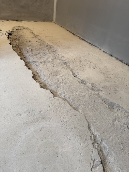Sprucken betonggolv med längsgående spricka, nära en grå vägg.