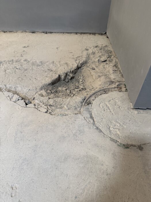 Sprucken betonggolv i ett hörn med synligt skadat underlag och rördetalj.