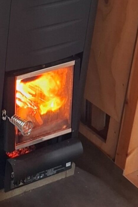 Braskamin med eldsflammor i glasluckan, installerad inomhus med träpanelväggar.